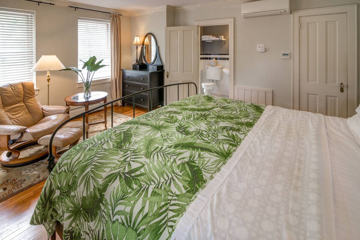 Une chambre à coucher comprenant un lit, une chaise, une commode, un miroir et une salle de bain visible à travers une porte ouverte.