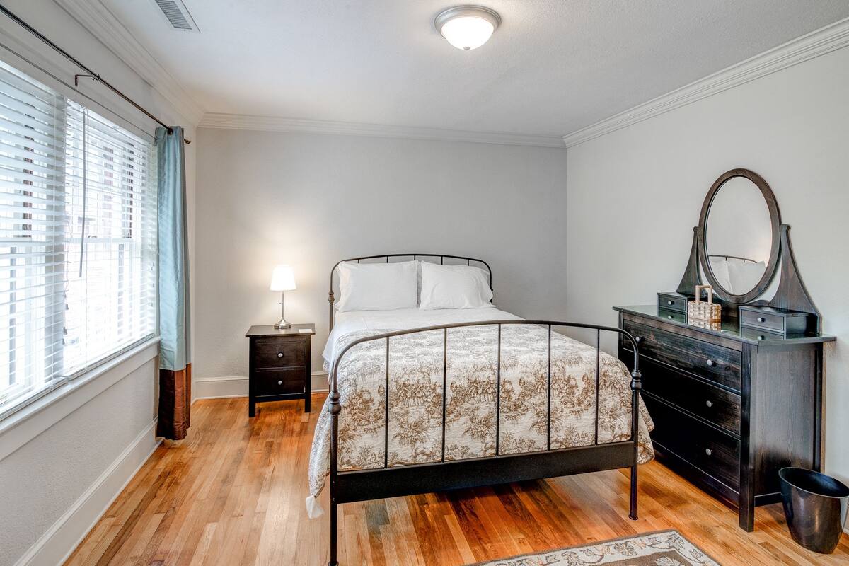 Une chambre à coucher comprenant un lit, une table de chevet, une commode, un miroir et une fenêtre avec des stores.