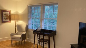 Un salon avec un plancher en bois, un fauteuil blanc, une table en bois foncé avec une chaise, une grande fenêtre avec des stores, un tableau encadré au mur et un téléviseur posé sur un meuble.