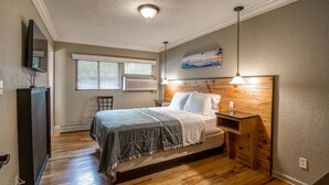 Une chambre avec un plancher en bois, un lit avec une tête de lit, une table de chevet, une commode, une fenêtre avec des stores et un tableau accroché au mur.
