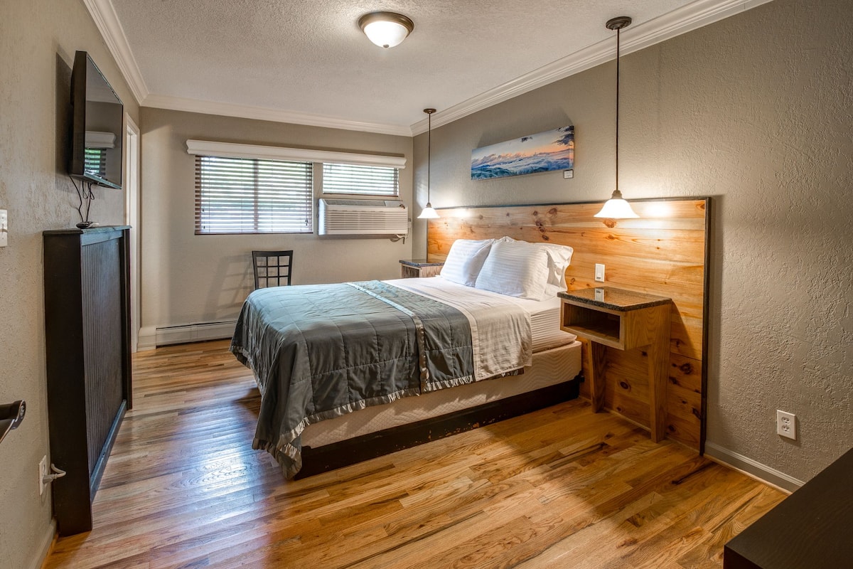 Une chambre avec un plancher en bois, un lit avec une tête de lit, une table de chevet, une commode, une fenêtre avec des stores et un tableau accroché au mur.
