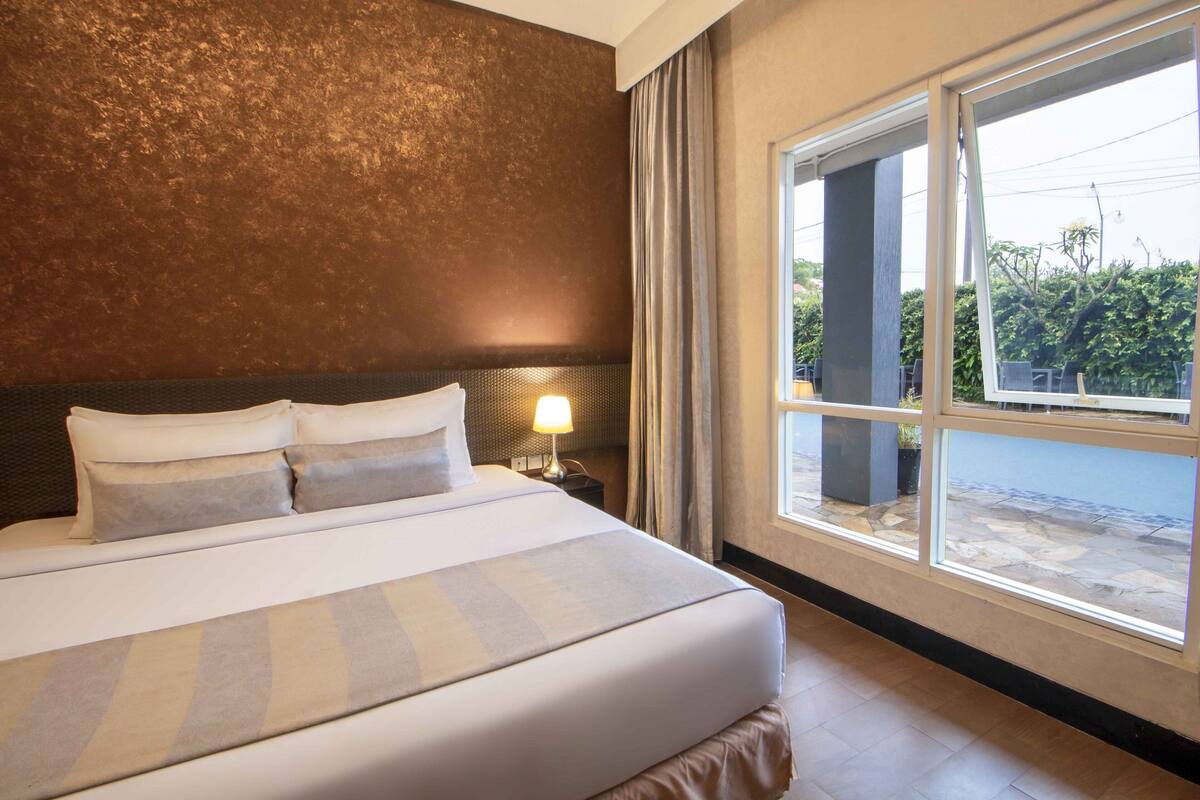Een hotelkamer met een groot bed, een nachtkastje met lamp en uitzicht op het omliggende landschap door een groot raam.