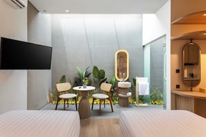 Quarto de hotel moderno com televisão de tela plana, duas camas, uma pequena área de refeições com cadeiras, pia e espelho.