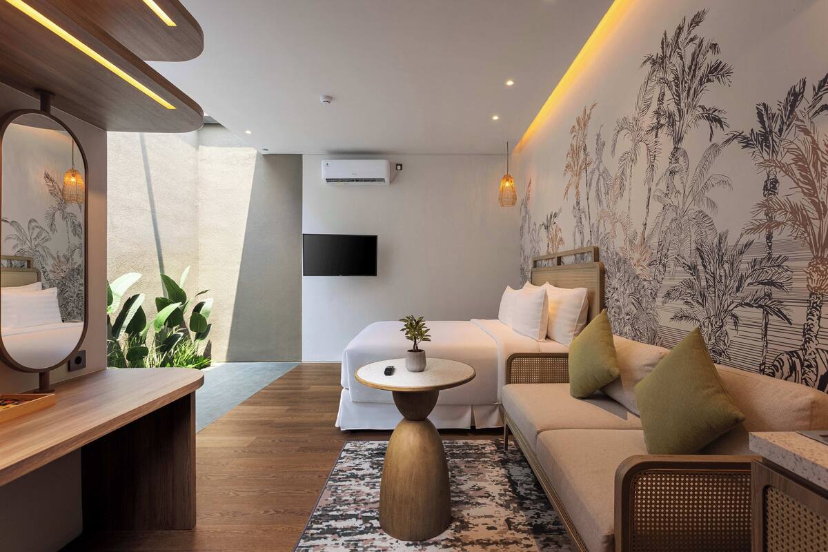 Een moderne hotelkamer met een bank, een ronde salontafel, een grote spiegel, een flatscreen televisie en een muur met een plantenmural in tropische stijl.