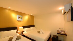A hotel room with two beds, a bedside table with a lamp, a small desk with a chair, and an air conditioning unit on the wall.