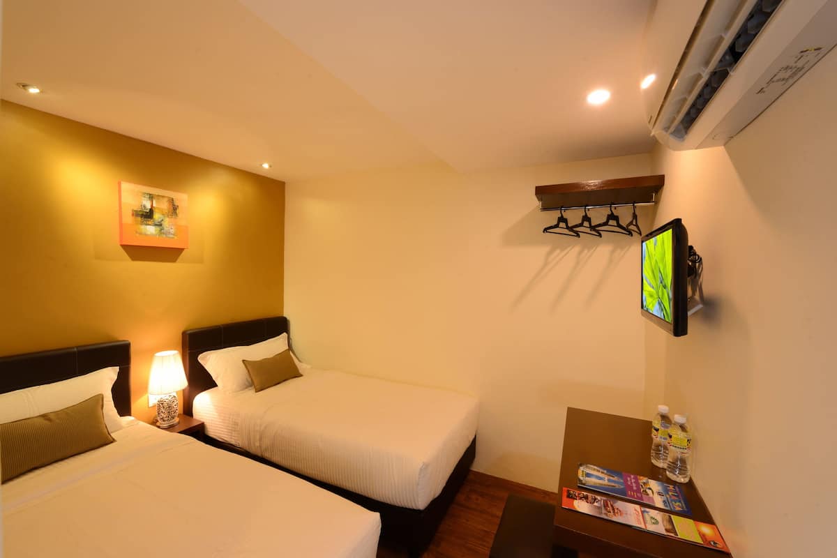 Ein Hotelzimmer mit zwei Betten, einem an der Wand montierten Fernseher, einem kleinen Tisch mit Wasserflaschen und Broschüren sowie einer Klimaanlage.