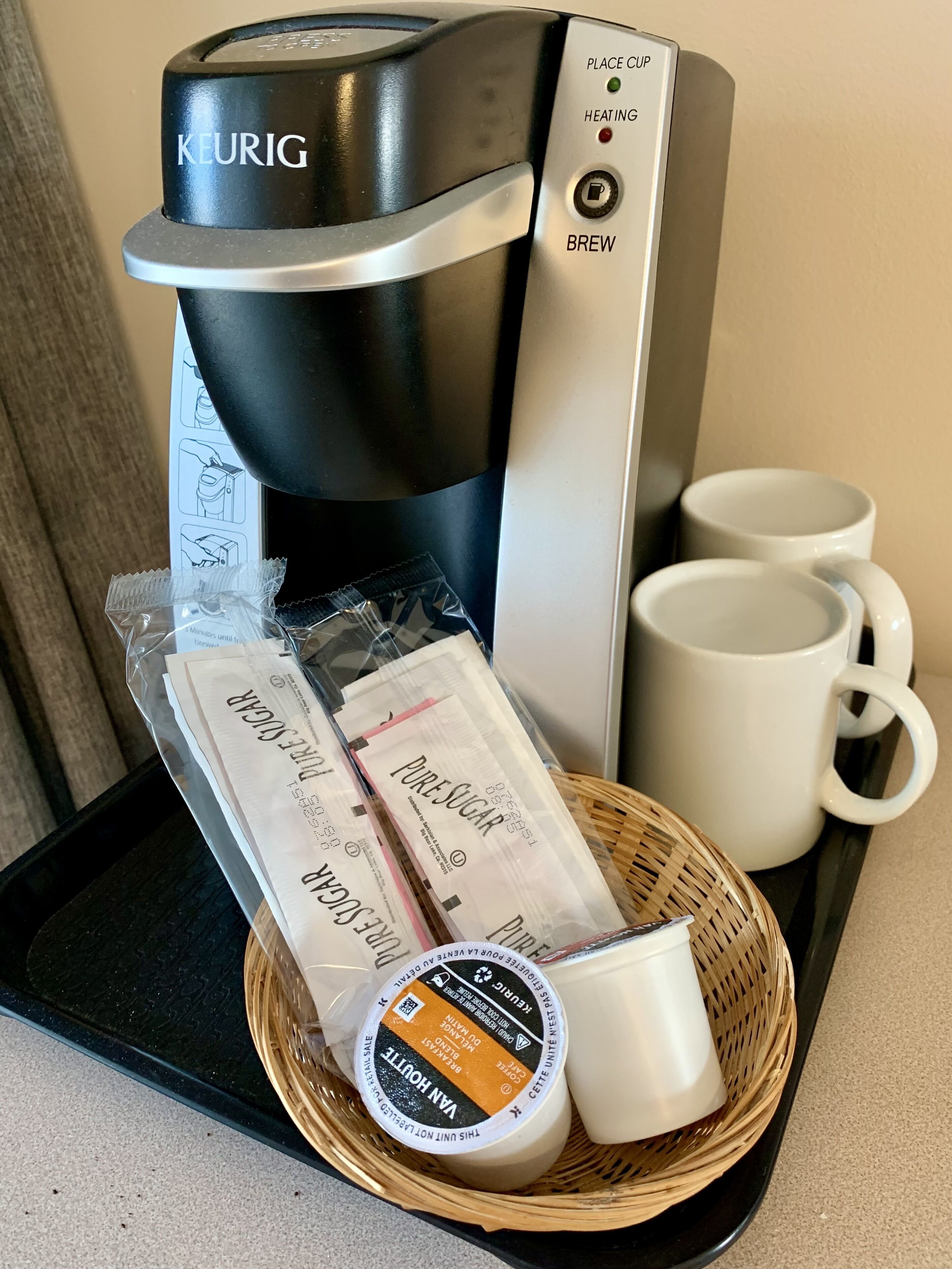 A Keurig coffee maker with a cup of coffee, two mugs, and a basket containing a coffee pod, a coffee bag, and a small container of cream.
