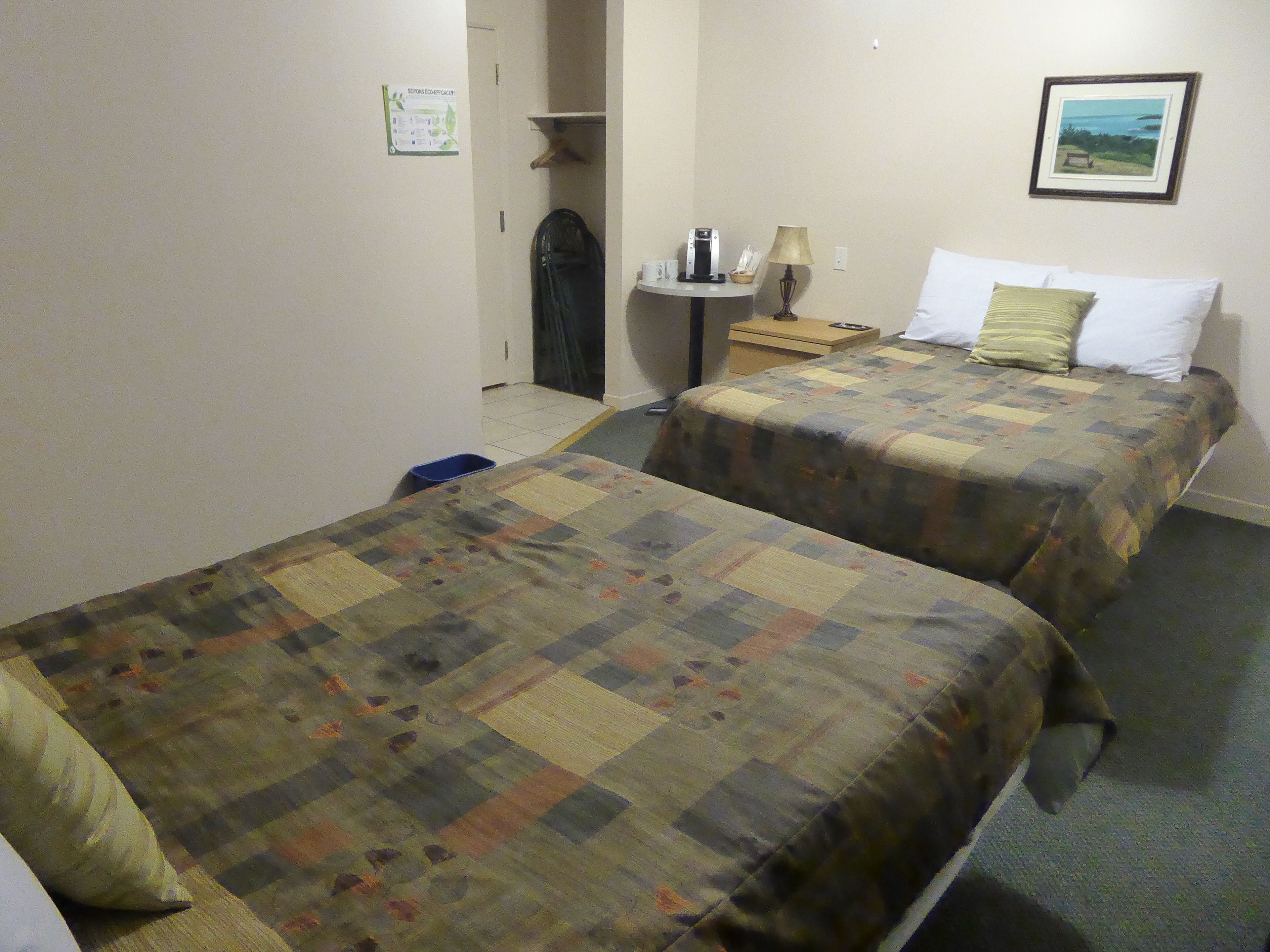 Two beds with checkered bedding, a nightstand with a lamp, a small table with a coffee maker, and a framed picture on the wall.