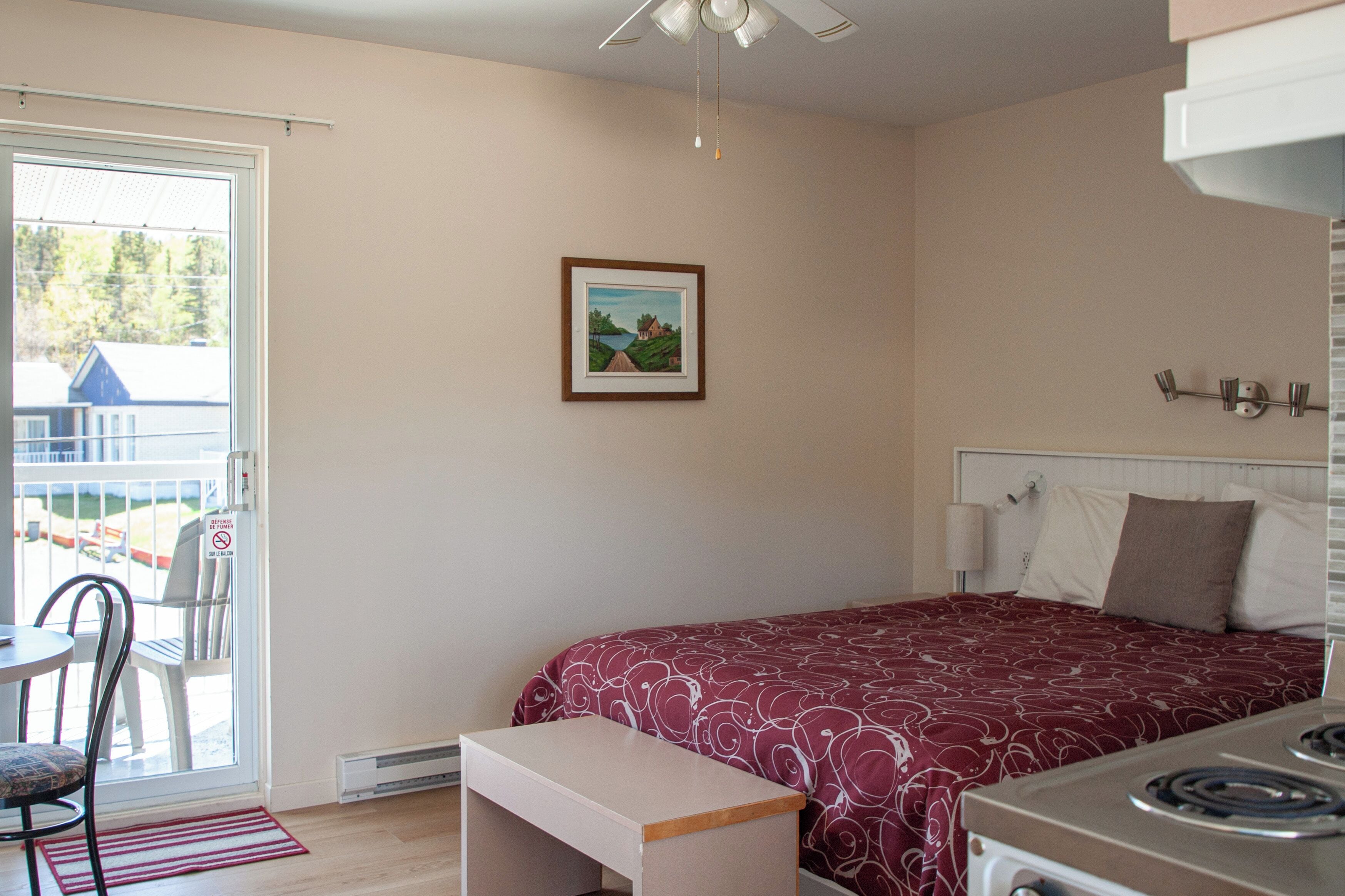 Un dormitorio con cama, una mesita, un banco, un ventilador de techo, una estufa y un cuadro en la pared.