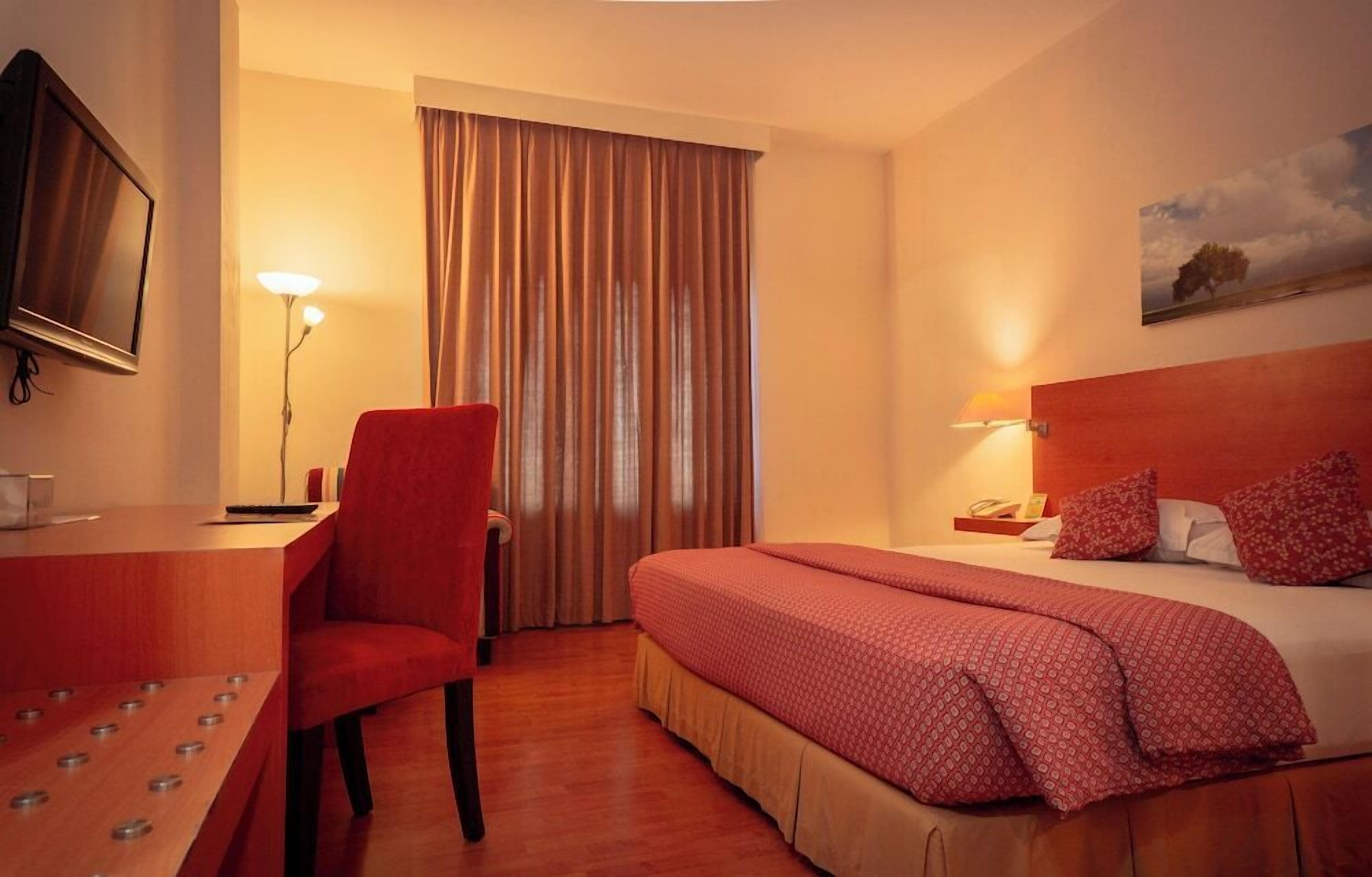 A hotel room with a bed, a desk, a chair, a TV, and a painting on the wall.