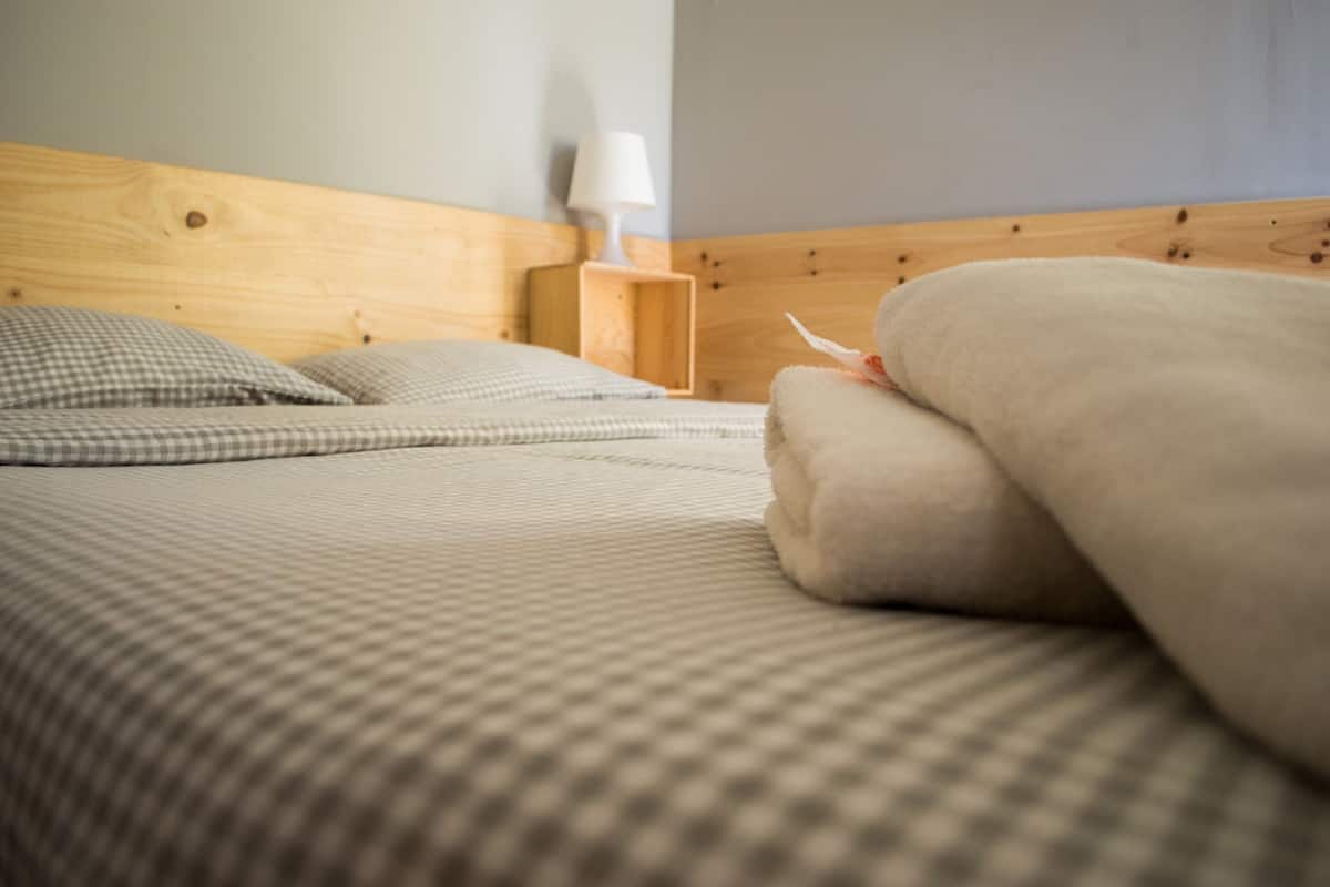 Un lit bien fait, avec du linge de lit à carreaux et uni, une tête de lit en bois et une table de chevet avec une lampe.