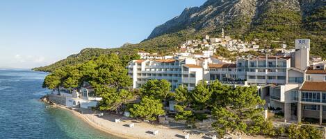 Un hotel frente al mar con varios pisos, una playa en primer plano y montañas al fondo.
