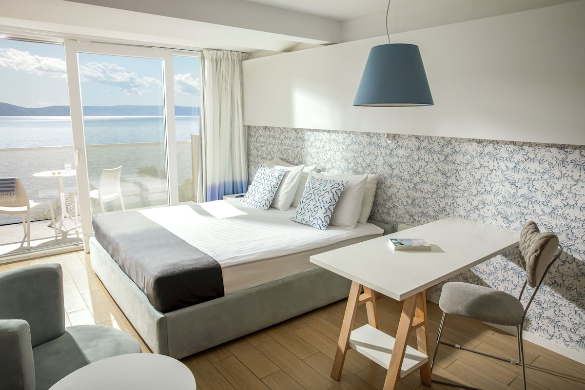 A modern hotel room with a large bed, a balcony with a view of the sea, a small table, and a chair.