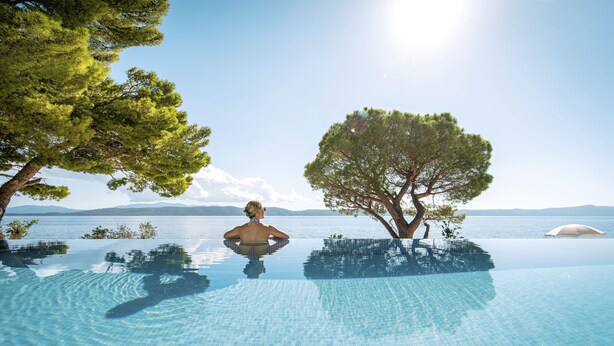 A person relaxing in a clear infinity pool with a view of the sea and mountains.
