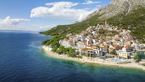 A coastal town with buildings along the shoreline, a clear view of the sea, and a hilly backdrop.