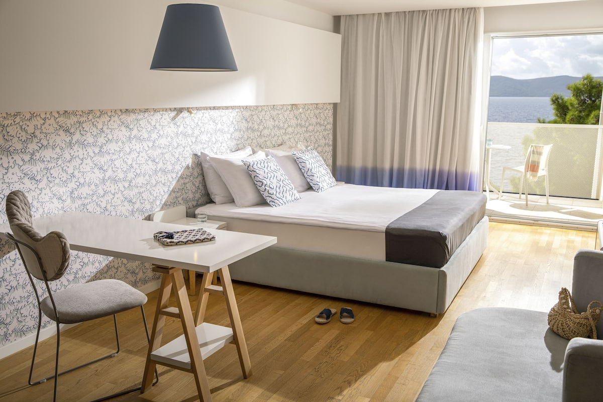 A modern hotel room with a bed, a table, a chair, and a view of the sea.