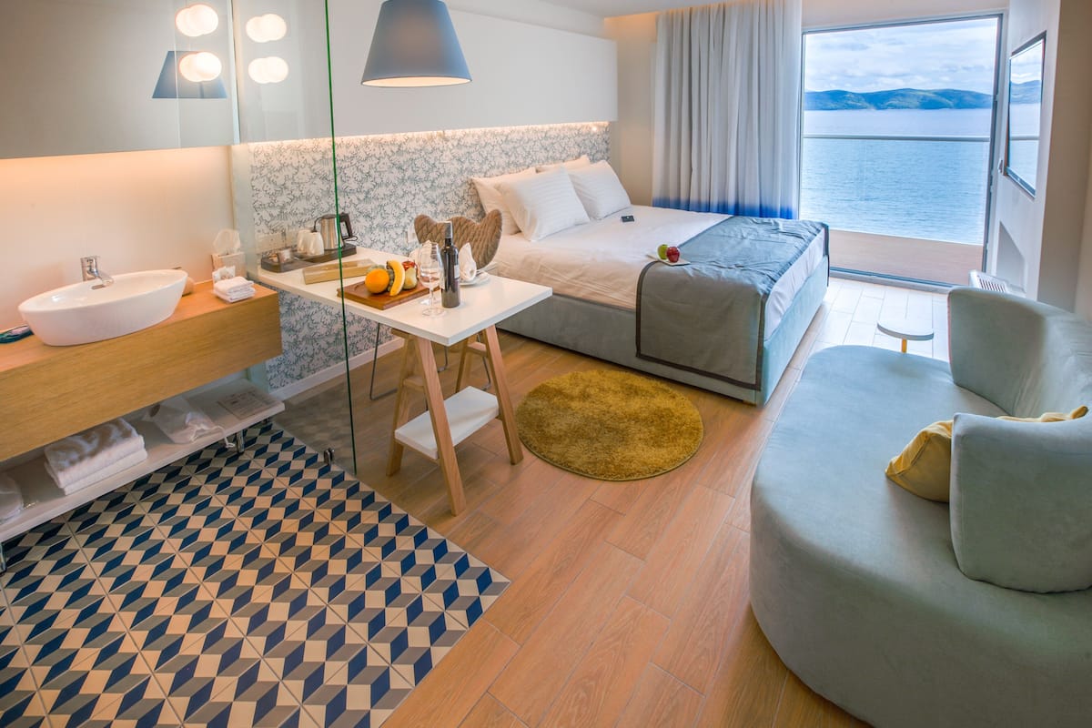 A modern hotel room with a large bed, a desk with a sink, a small table with fruits and a bottle, a round rug, and a view of the sea.