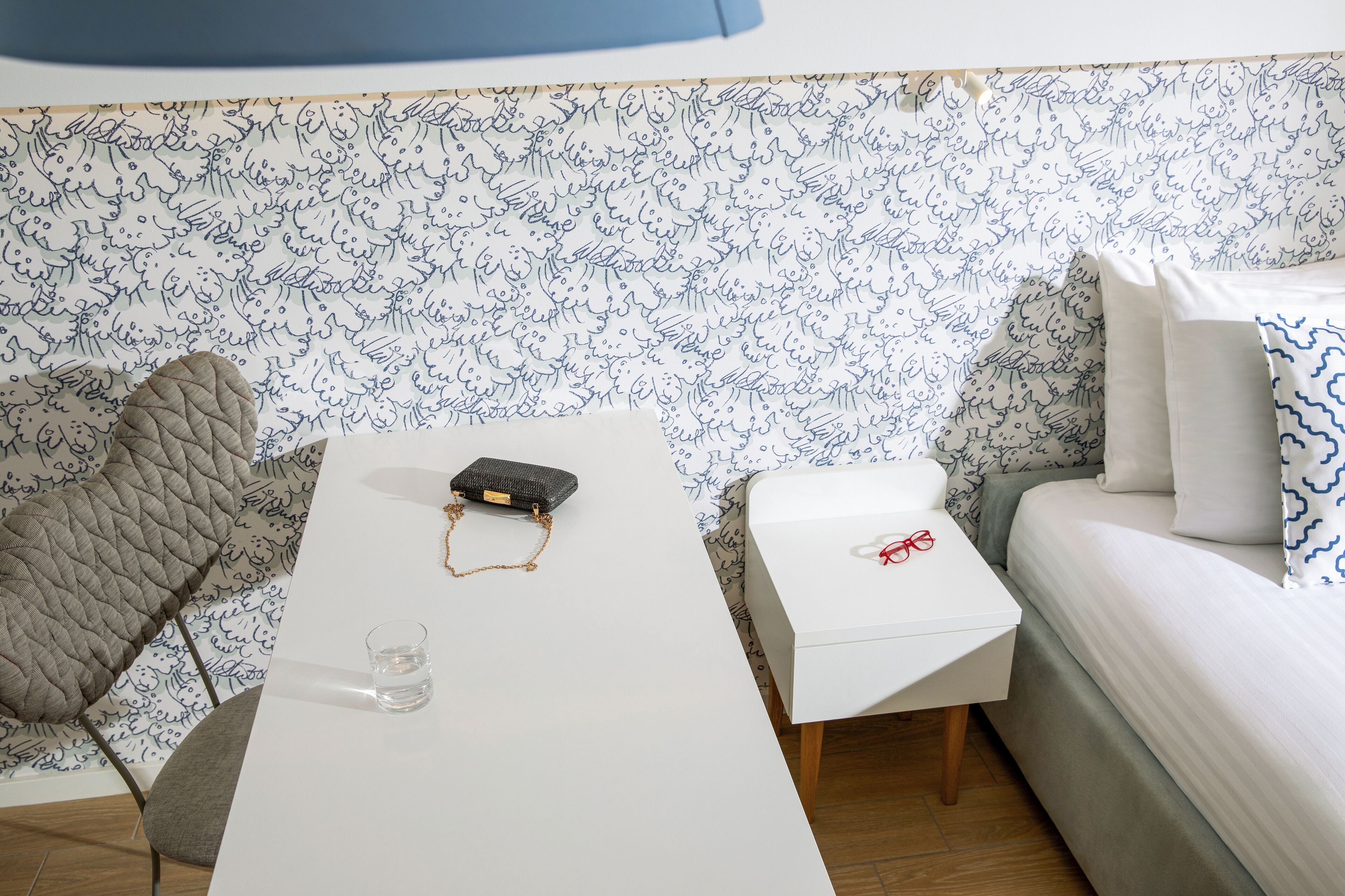Ein Zimmer mit einem weißen Tisch, einem kleinen weißen Nachttisch, einem grau gepolsterten Sessel, einem Bett mit Kissen in Weiß und Blau und einer Wand mit einem blau-weißen abstrakten Muster.