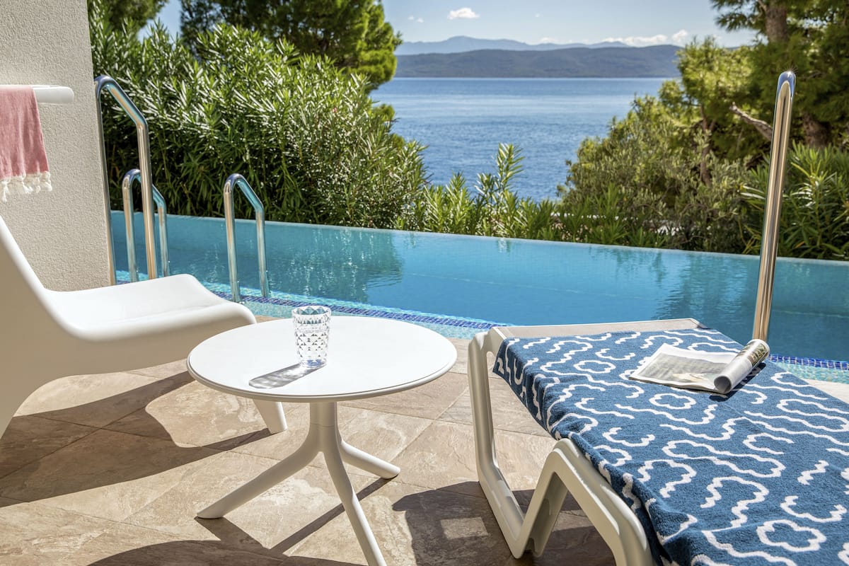 A poolside area with a view of the sea, a lounge chair, a small table, and a towel.
