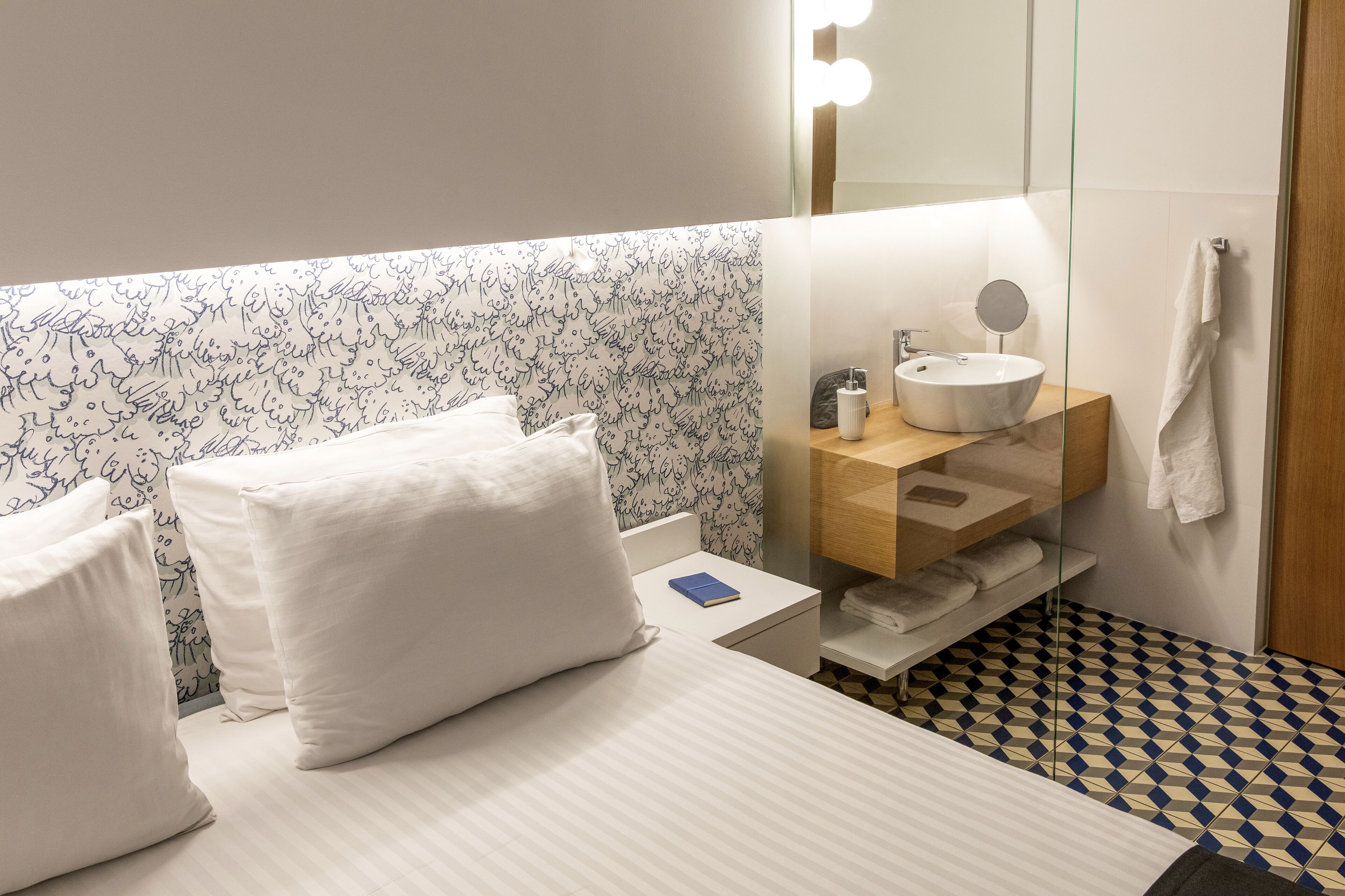 Habitación de hotel con cama, almohadas blancas, mesita de noche con un libro, baño con lavamanos, espejo y toallero.
