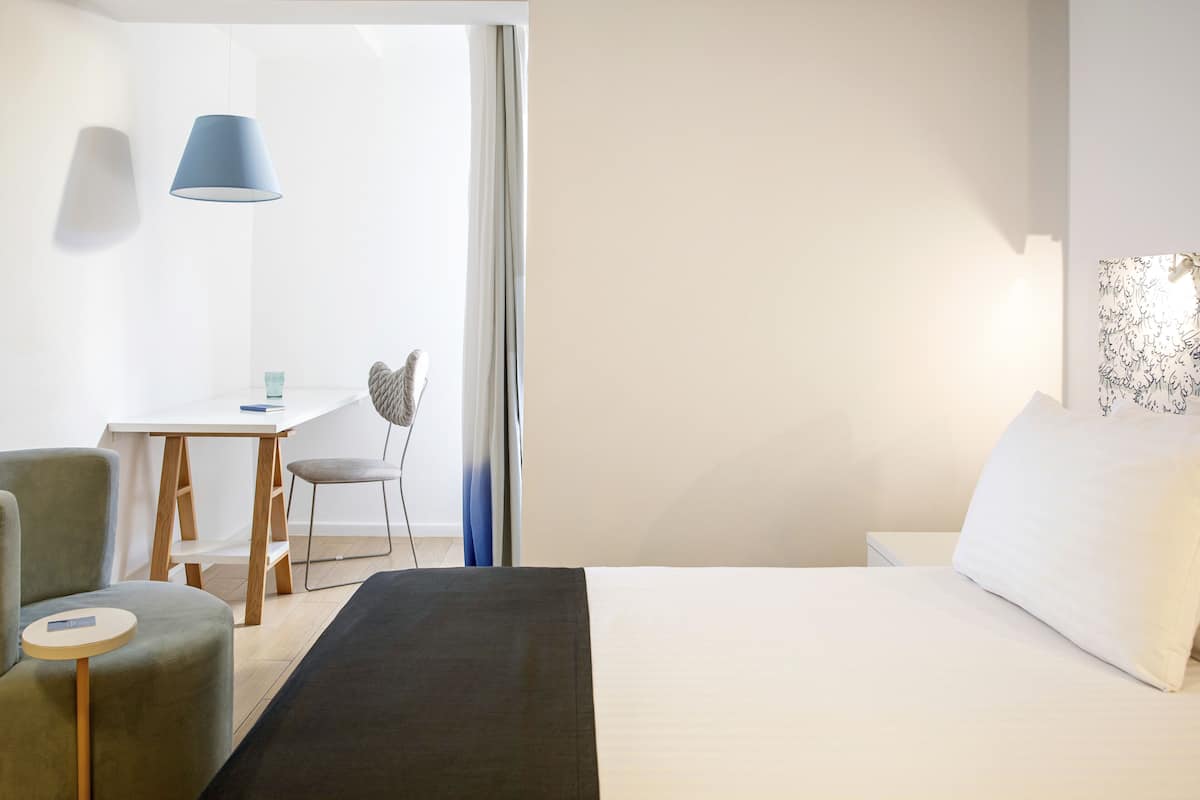 A hotel room with a bed, a desk, a chair, and a lamp.