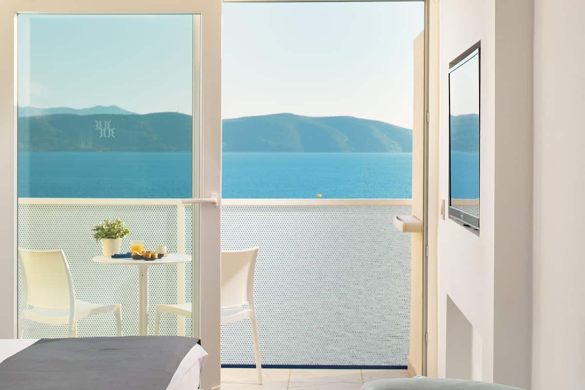 A hotel room with a bed, a round table with fruit and a drink, and a balcony with a view of the sea.