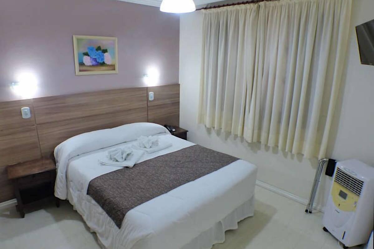 A hotel room with a bed, a bedside table, a lamp, a painting on the wall, and a window with curtains.