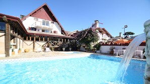 A swimming pool with a waterfall feature, surrounded by a building with a red roof and white walls. - Hotel JB (Campos do Jordão)