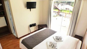 A hotel room with a bed, a flat-screen TV mounted on the wall, a window with curtains, and a view of the outside. - Hotel JB (Campos do Jordão)