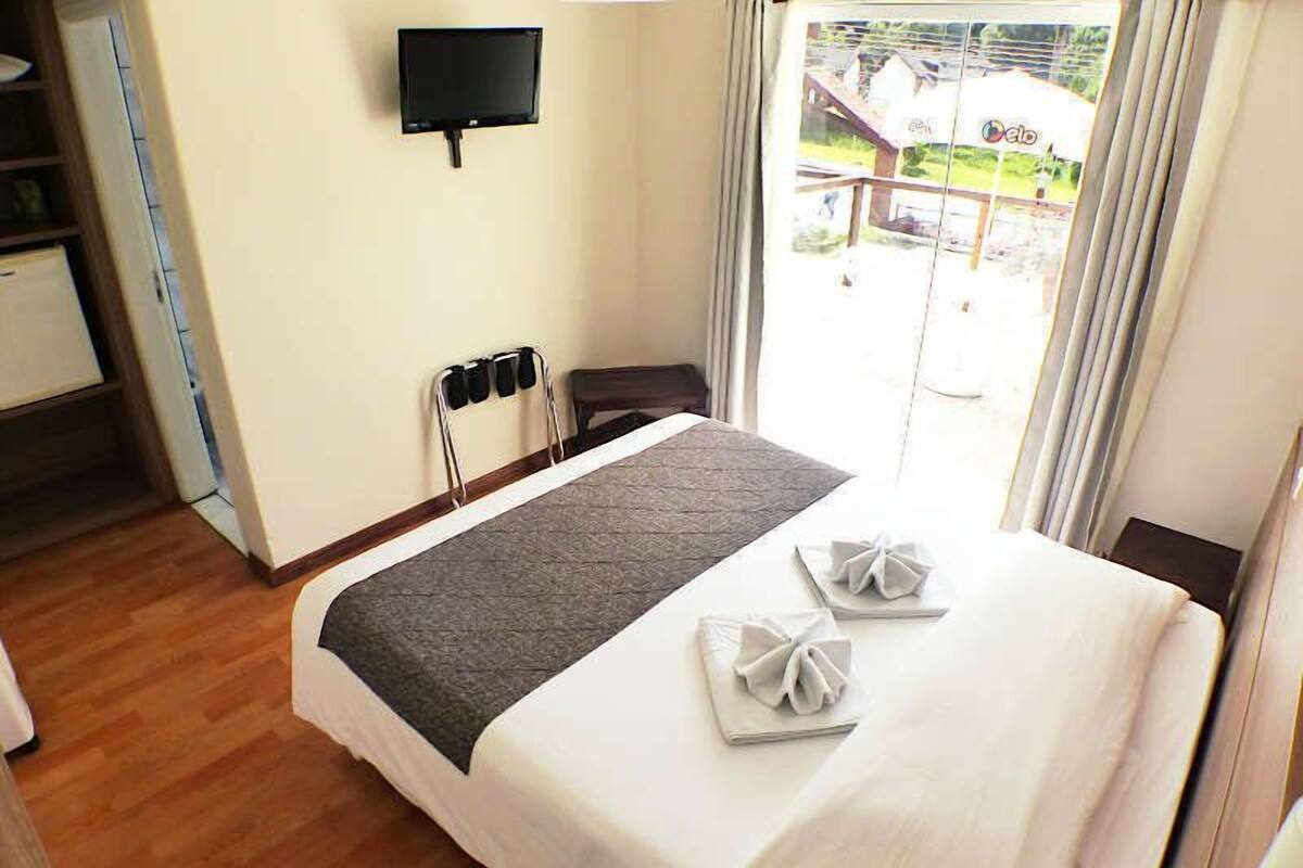 A hotel room with a bed, a flat-screen TV mounted on the wall, a window with curtains, and a view of the outside.