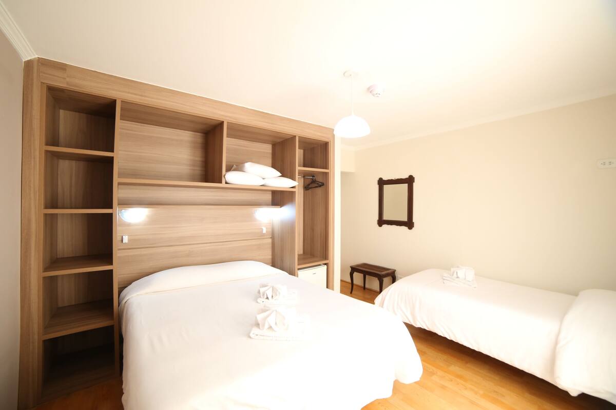 A hotel room with two beds, a wooden wardrobe, a small table, and a mirror on the wall.