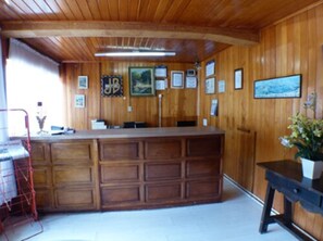Un interior de madera con mostrador de recepción, cuadros enmarcados en la pared y una planta en maceta.