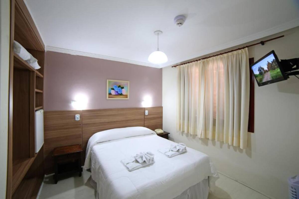 A hotel room with a bed, bedside table, lamp, TV, and a painting on the wall.