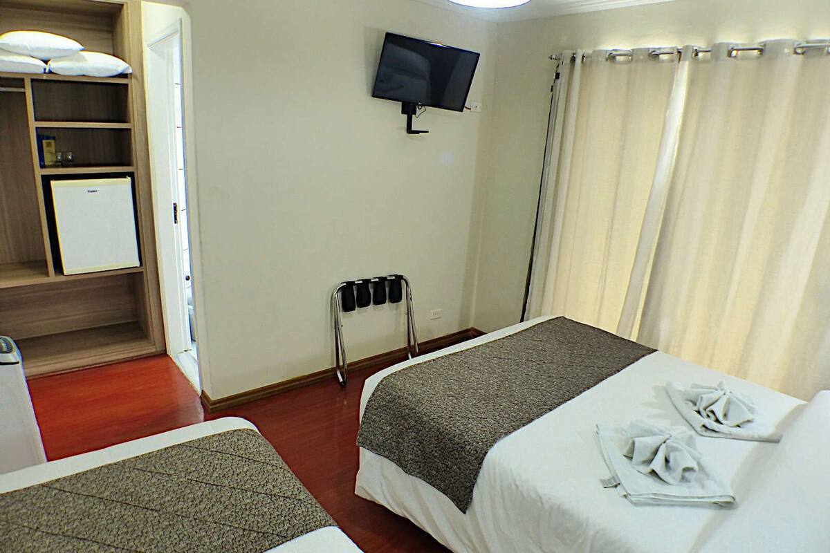 A hotel room with two beds, a TV mounted on the wall, a small kitchenette, and a window with curtains.