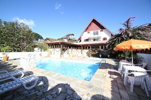 A swimming pool area with lounge chairs, a stone wall, and a building with a red roof. - Hotel JB (Campos do Jordão)
