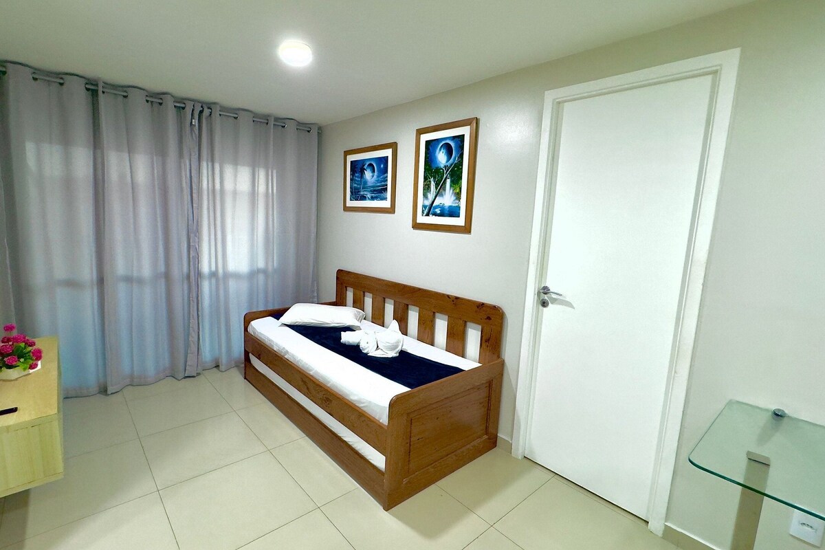 A bedroom with a bed, a bedside table with a vase of flowers, a door, and two framed pictures on the wall.