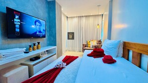 Una habitación de hotel moderna con una televisión de pantalla plana mostrando una película, una cama bien hecha con almohadas rojas, una pequeña zona de estar con un sillón, y un estante con artículos de aseo.