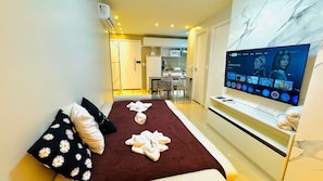 Une chambre d’hôtel avec une télévision, un lit avec des serviettes, un canapé avec des coussins à motifs, un coin cuisine et un mur en marbre.