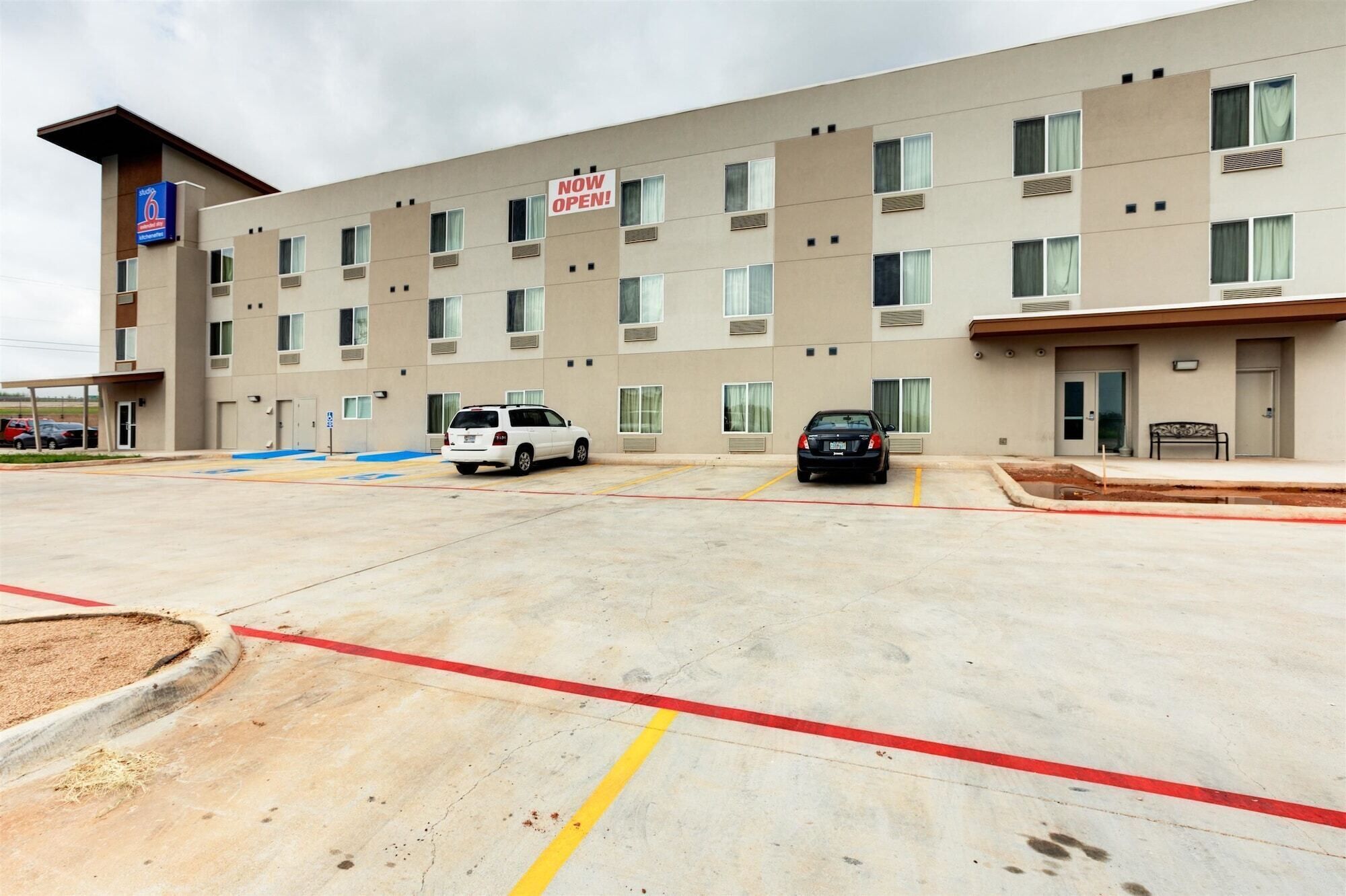 A multi-story hotel with a 'NOW OPEN' sign, a parking lot with marked spaces, and a building entrance.