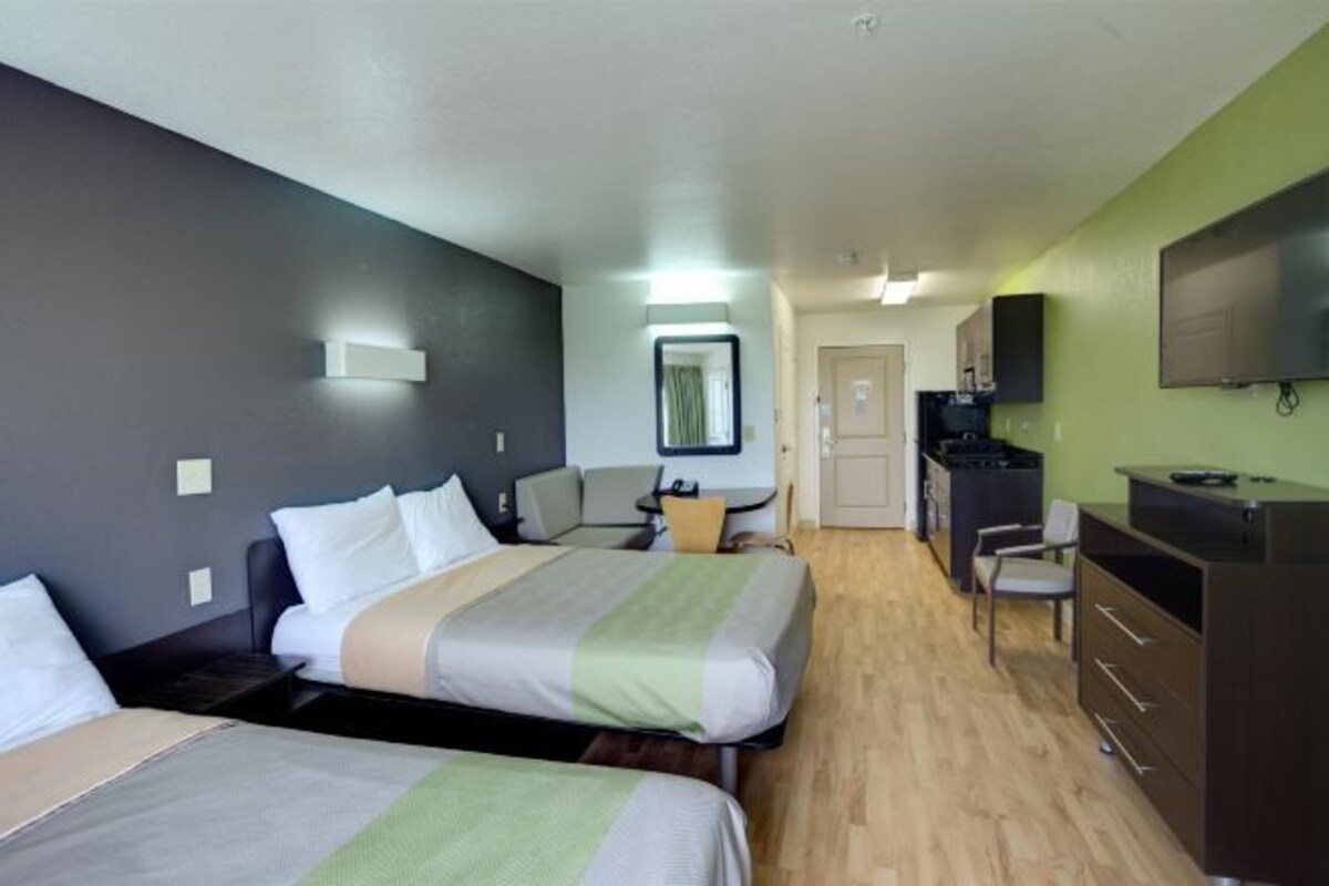 Quarto de hotel com duas camas, uma mesa, uma cadeira, uma televisão e uma kitchenette pequena.