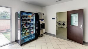 A room with a vending machine stocked with beverages, a guest laundry area with washing machines, and a door leading to another room. - Studio 6 Sweetwater, TX (Sweetwater)