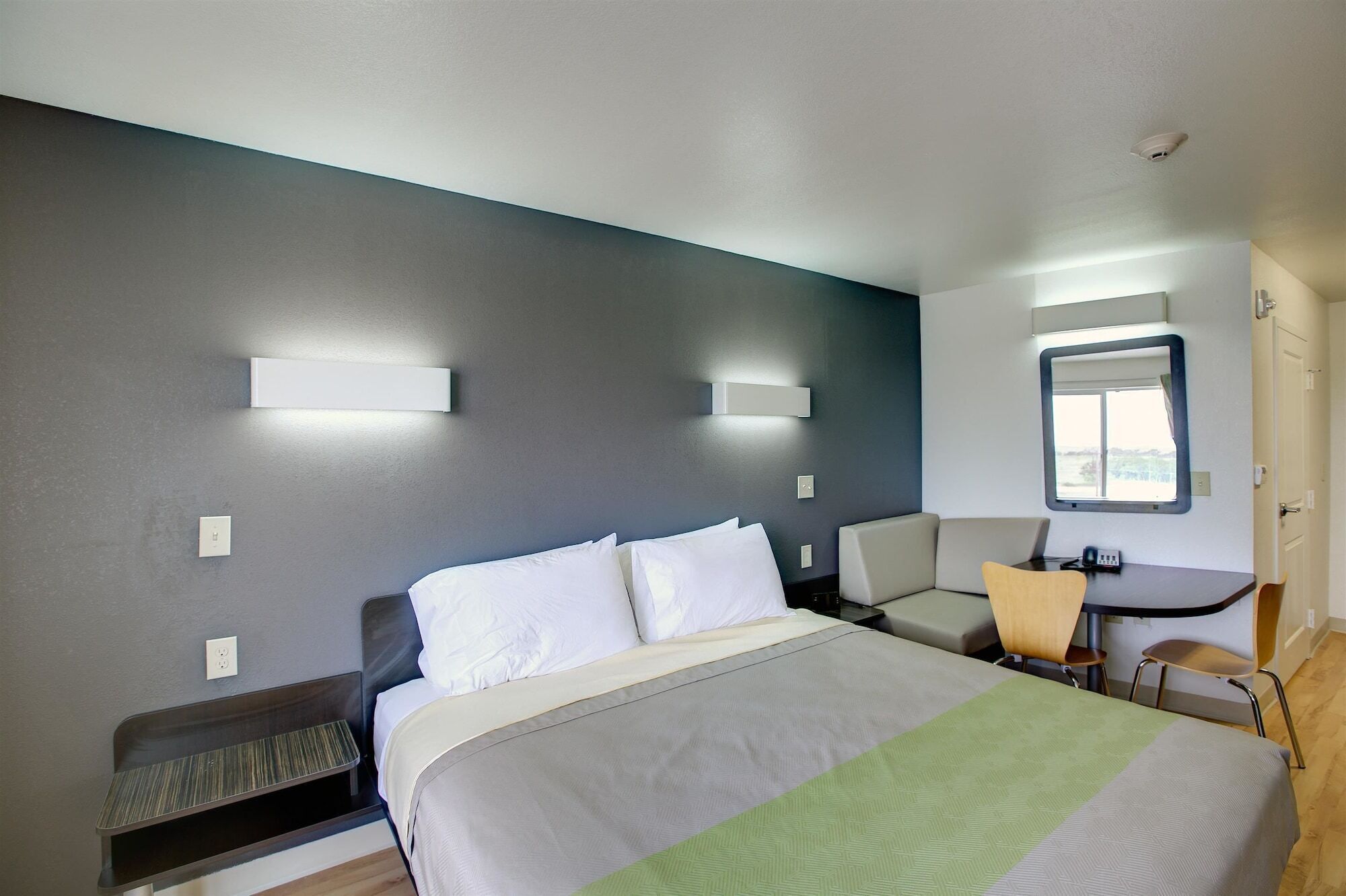 A modern hotel room with a bed, bedside table, chair, and a small desk with a chair.