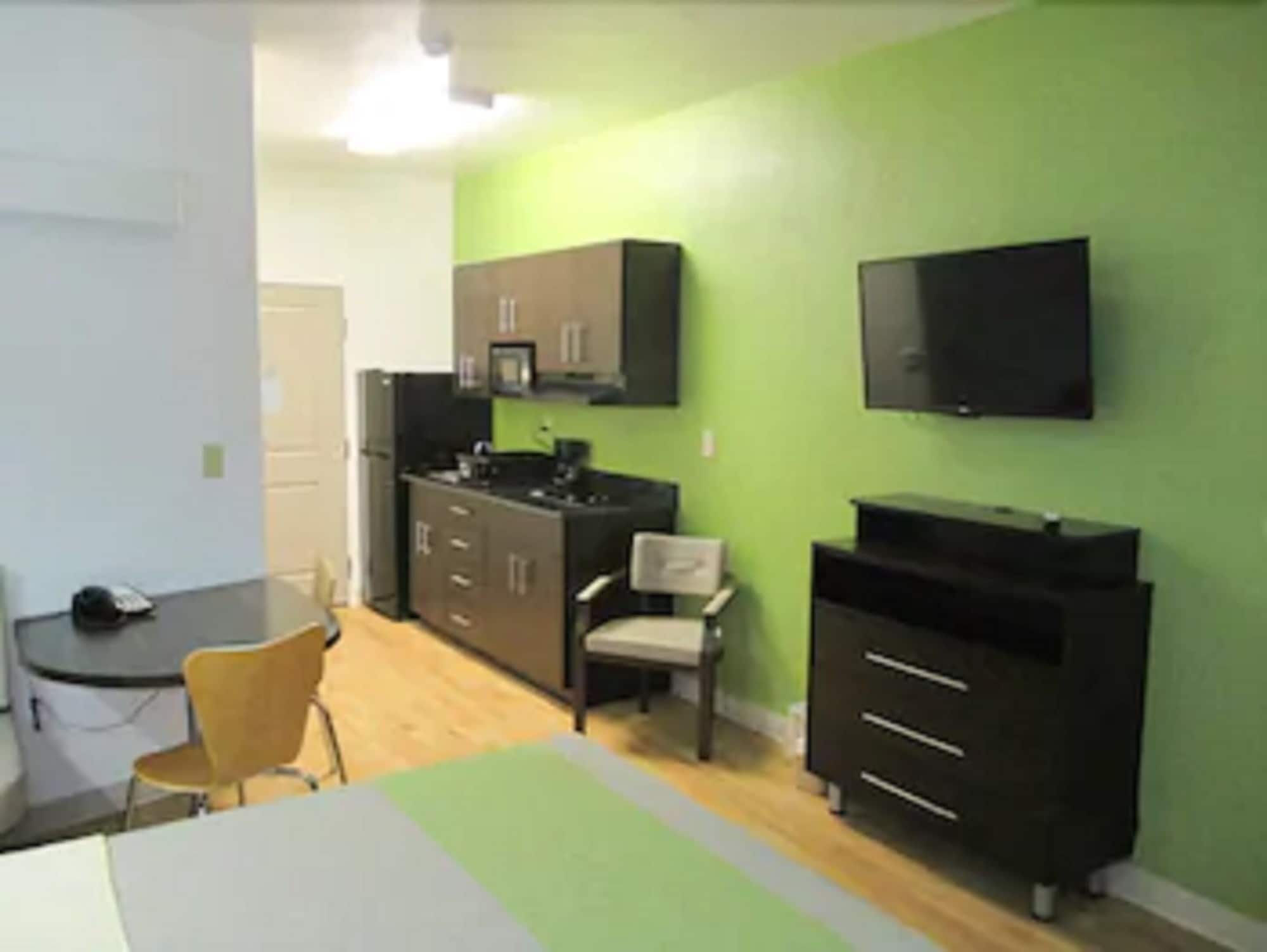 A compact kitchen with a refrigerator, microwave, and cabinets. There is a small dining area with a chair and a table. A flat-screen TV is mounted on the wall.