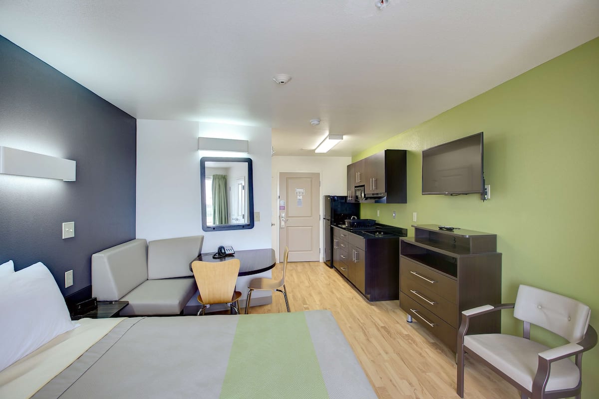 A modern hotel room with a bed, a seating area, a kitchenette, and a TV mounted on the wall.