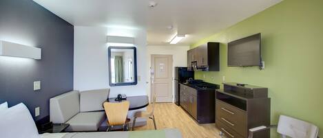 A modern hotel room with a bed, a seating area, a kitchenette, and a TV mounted on the wall.