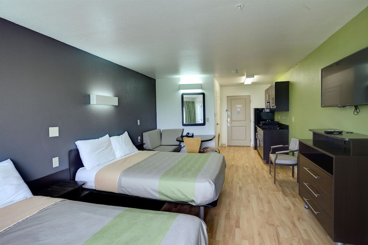A hotel room with two beds, a desk, a chair, a kitchenette, and a television.