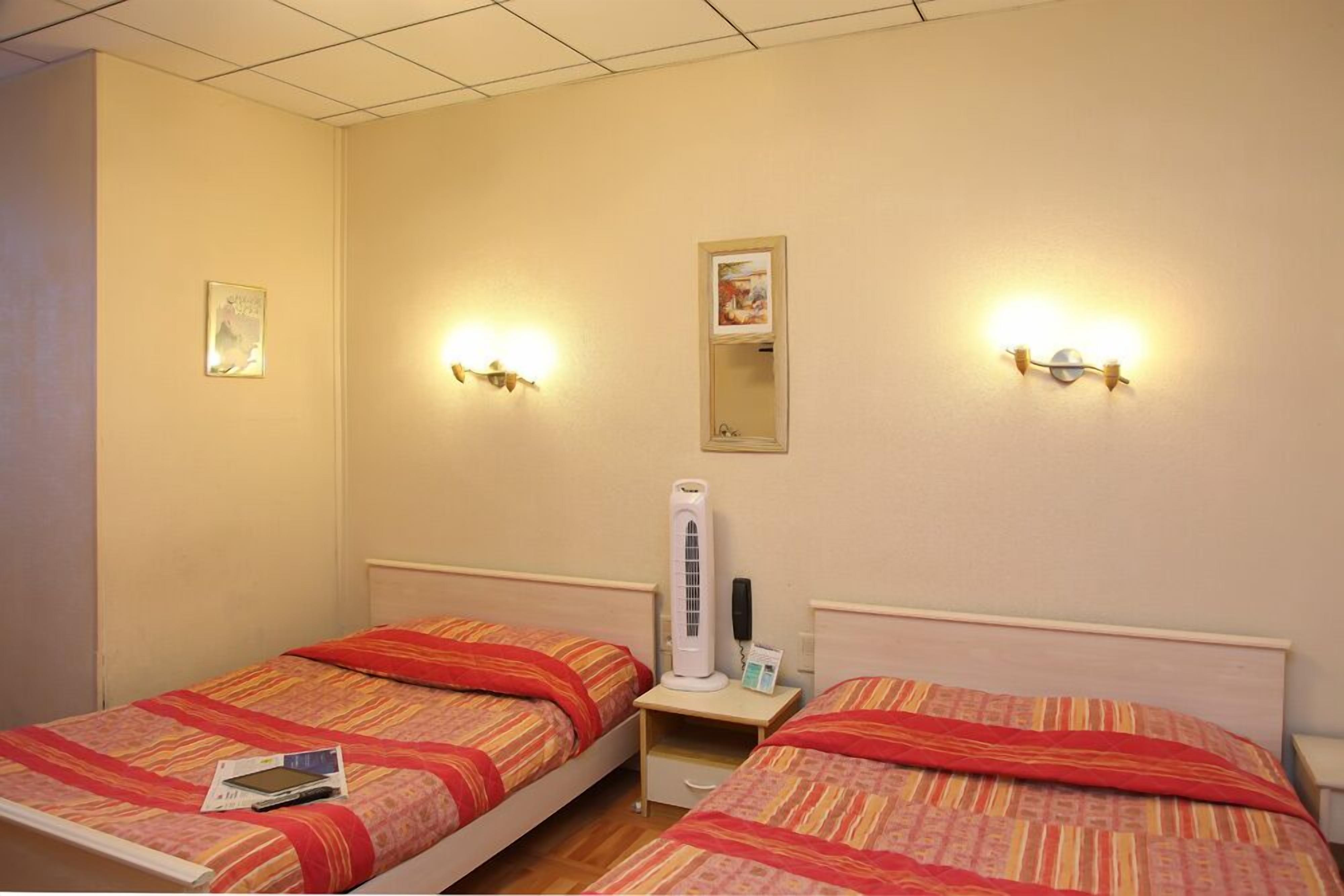 A hotel room with two single beds, each with a red and orange striped bedspread, a bedside table with a lamp, a wall-mounted mirror, and a wall-mounted light fixture.