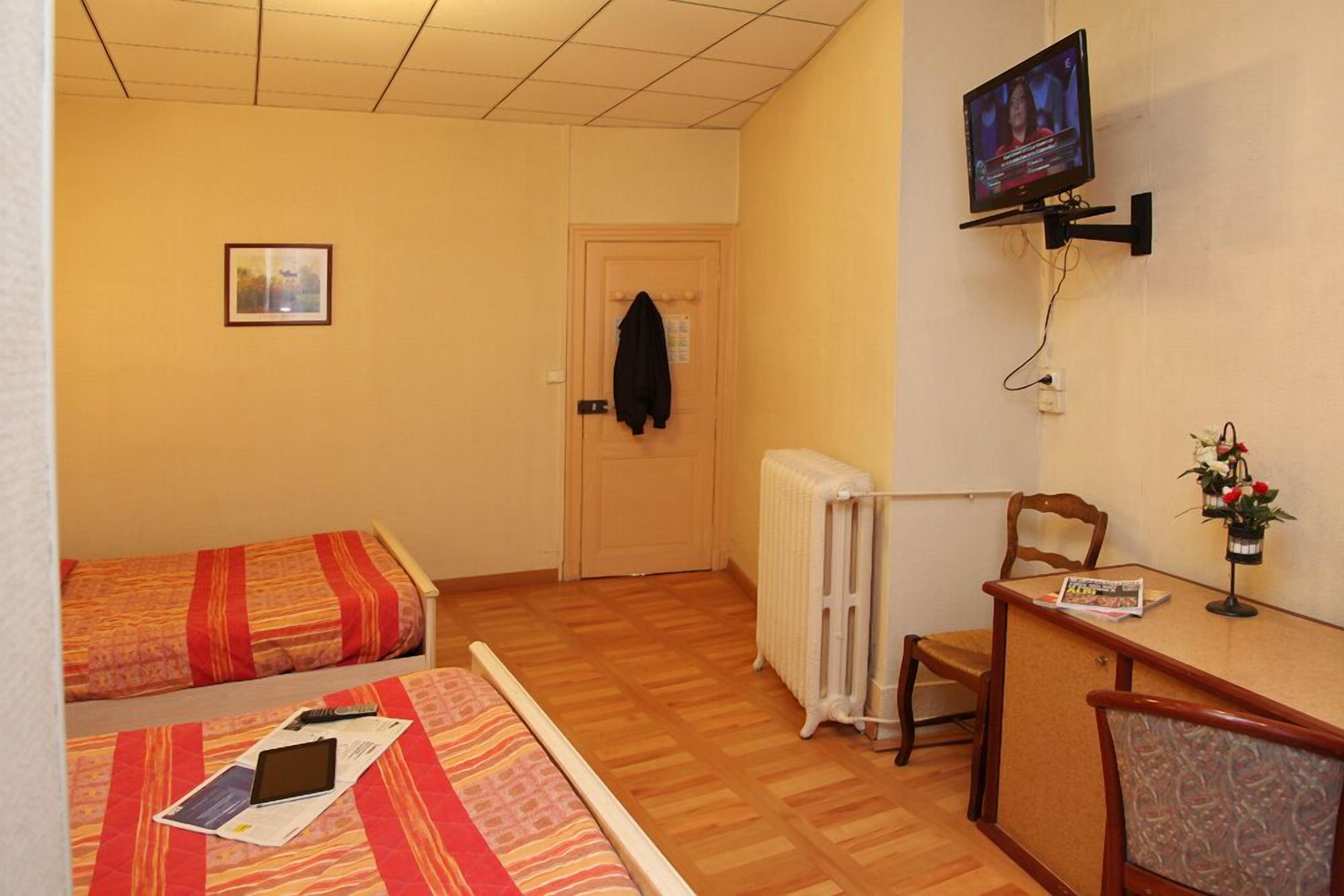 A small hotel room with two single beds, a TV mounted on the wall, a radiator, a desk with a chair, and a small table with a vase of flowers.