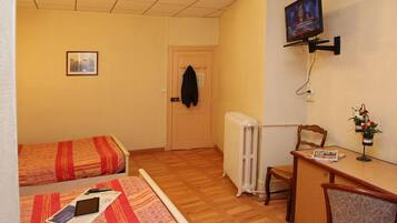 A small hotel room with two single beds, a TV mounted on the wall, a radiator, a desk with a chair, and a small table with a vase of flowers.