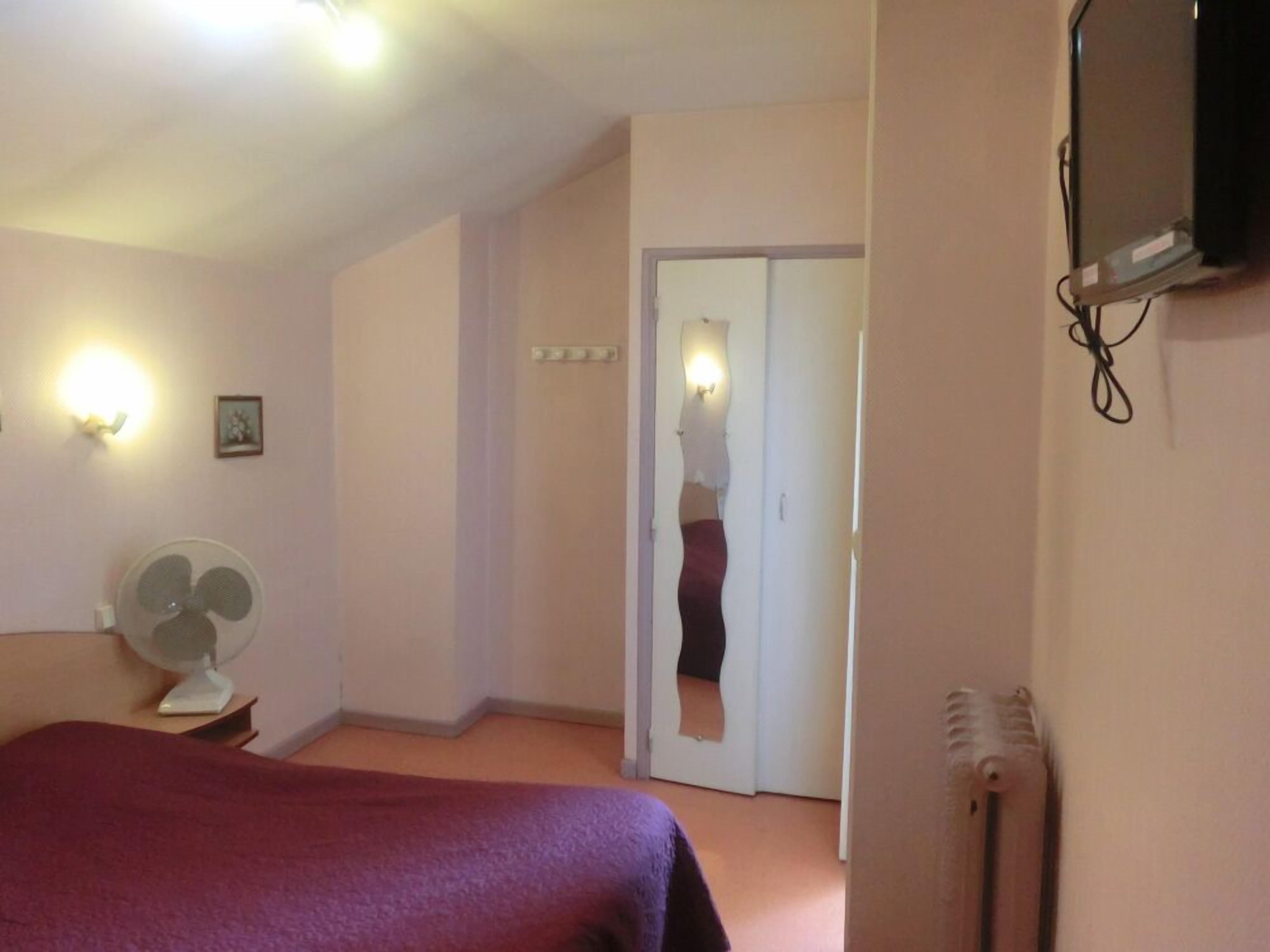Une chambre d’hôtel équipée d’un lit, d’un ventilateur sur pied, d’un téléviseur fixé au mur et d’une porte donnant sur une autre pièce.